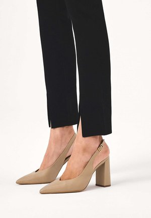 Picioare inferioare purtând pantaloni negri scurtați cu fante laterale, asortați cu pantofi bej cu toc gros, cu partea din spate deschisă și barete cu cataramă.