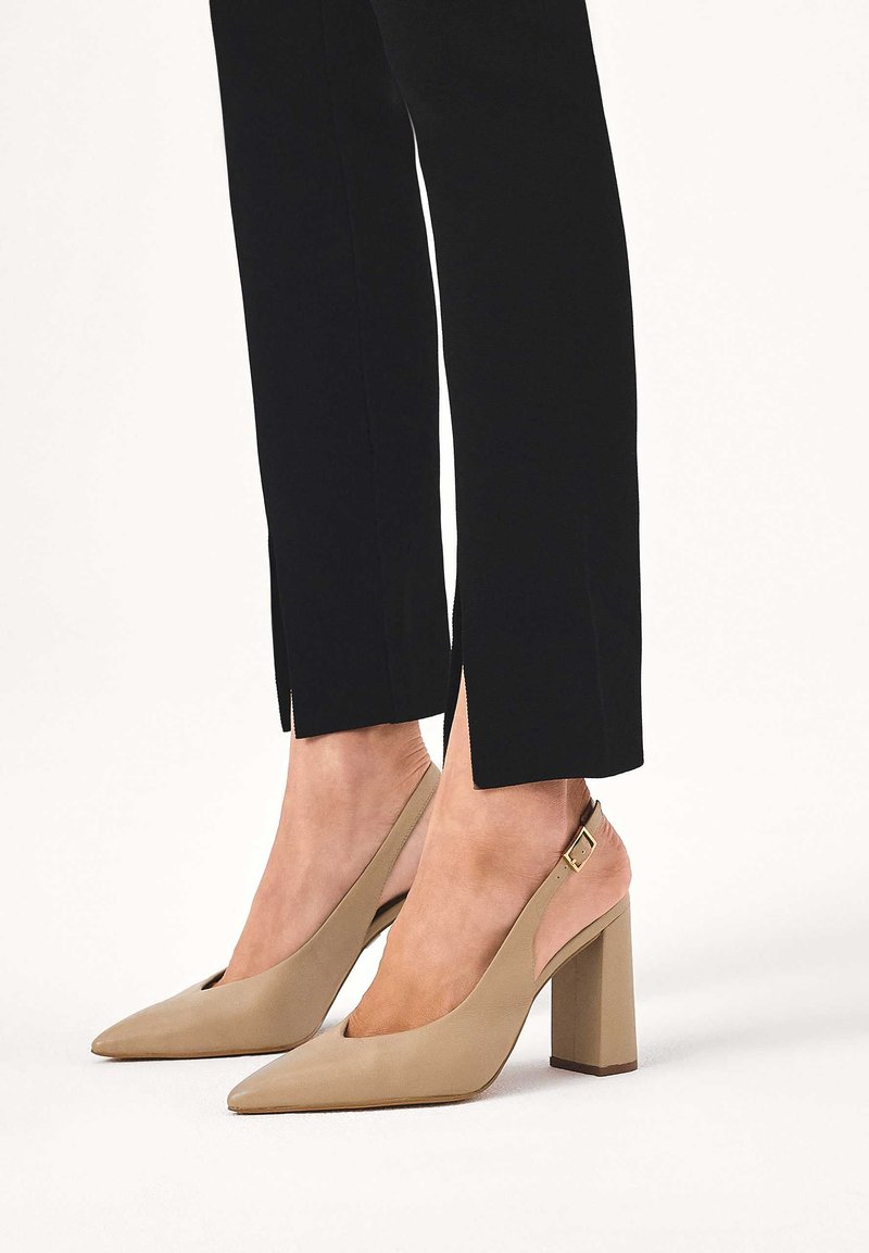 Untere Beine in schwarzen, gekürzten Hosen mit seitlichen Schlitzen, kombiniert mit beigen, spitz zulaufenden Slingback-Pumps mit Blockabsatz und Schnallenriemen.