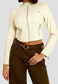 Woman wearing a cropped cream leather jacket with front zippers and high-waisted brown pants with a brown belt featuring a gold buckle.