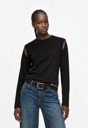 Femme aux cheveux tressés porte un haut noir à manches longues avec des découpes aux épaules, un jean bleu et une ceinture noire avec une boucle argentée et des clous.