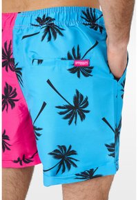 Homme portant un short de bain bleu et rose avec des motifs de palmiers noirs et une petite étiquette rose portant l'inscription Opposuits.