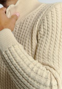 Beige gebreide trui met geribbelde kraag en manchetten. Heeft een gestructureerd, kabelgebreid patroon. Close-up van de stof en een hand die de kraag vastgrijpt.