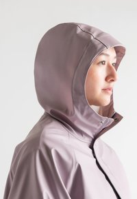 Giacca impermeabile rosa chiaro con cappuccio aderente, chiusura frontale con cerniera e bottoni a pressione. Texture liscia con cuciture minime per un design elegante.