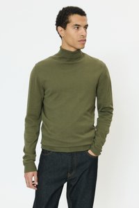 Olivno zelena pulover z visokim ovratnikom iz mehkega blaga z dolgimi rokavi in rebrastimi manšetami, nošen z temno modrimi kavbojkami. Gladek tekstura, oprijet dizajn.