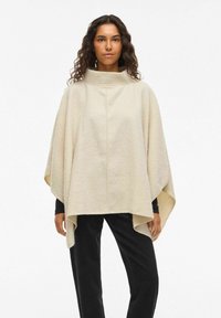 Beige uld poncho med høj krave, der har en løs pasform og asymmetrisk kant. Blød tekstur, minimale syningsdetaljer.