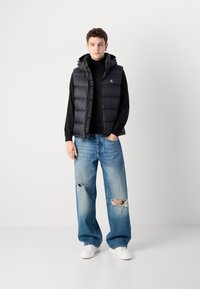 Zwarte puffer vest over een zwarte trui, gecombineerd met lichtblauwe versleten wijde jeans. Witte sneakers en een neutrale achtergrond.