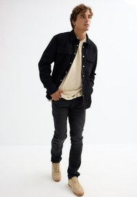 Camisa negra de botón con cuello, superpuesta a una camiseta beige clara. Jeans oscuros y zapatillas color beige completan el conjunto.