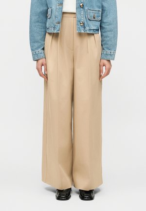 Μπεζ παντελόνι φαρδύ στο πόδι με πιέτες μπροστά, φορεμένο με cropped τζιν μπουφάν και μαύρα παπούτσια με τετράγωνη μύτη σε λευκό φόντο.