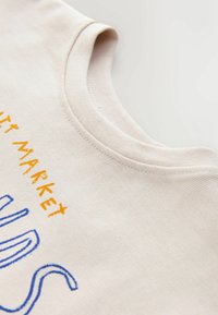 Világosbézs pamutpóló kerek nyakkivágással, kék és narancssárga hímzéssel, amelyen az "Fruits Market" felirat olvasható. Puha tapintású, hétköznapi kialakítás.