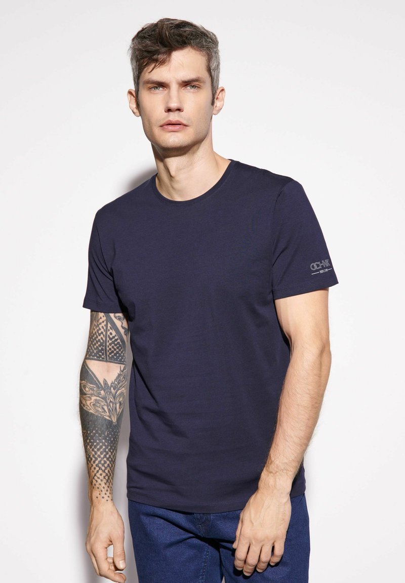 Granatowa bawełniana koszulka z okrągłym dekoltem, krótkimi rękawami i subtelnym logo na rękawie. Gładka faktura, klasyczny krój.