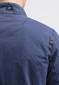 Mörkblå skjorta med en strukturerad krage, synliga sömdetaljer och en brun knappdekoration på axeln. Mjuk, texturerad tyg.