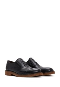Scarpe nere in pelle con finitura testurizzata, punta tonda e quattro fori per i lacci. Suola simile al legno con colore marrone a contrasto.