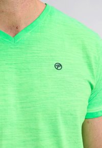Tricou scurt cu mâneci scurte de culoare verde deschis, de tip V, realizat dintr-un material ușor. Prezintă un logo mic, de culoare neagră, pe partea stângă a pieptului.