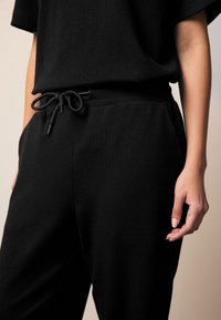 Juodi sportinės kelnės su elastine juosta ir raišteliais, pagamintos iš minkštos medžiagos. Turi šonines kišenes ir laisvą siluetą.