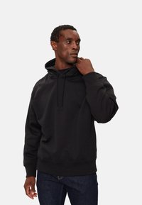Sudadera negra de algodón, con capucha ajustable, puños acanalados y un pequeño logo en el pecho. Llevada con vaqueros azules.