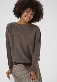 Brūns, vaļīgs adījuma džemperis ar plašu laivas izgriezumu un ribotām piedurknēm, kombinēts ar rakstainām svārkiem, kuriem ir pārbaudes dizains.