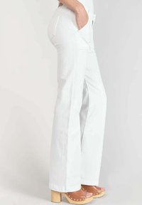 Jean flare blanc avec poches latérales, présentant des coutures contrastantes. Associé à des sandales à plateforme beiges ornées de clous décoratifs.