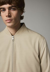 Beige bomberjakke med høj krave og frontal lynlås, med et glat, tekstureret stof og et minimalistisk design.