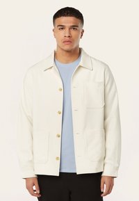 Witte button-up jas met twee voorzakken, lichtblauw binnenhemd. Zachte textuur, ontwerp met kraag en zichtbare stikseldetails.