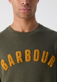 Tmavě zelená mikina s texturovaným, žlutým výšivkovým nápisem "BARBOUR" na přední straně. Kolo výstřih a měkká tkanina.