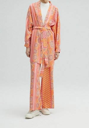 Person trägt einen orange-rosa Blumenmuster-Mantel mit Gürtel und passende weit geschnittene Hosen, kombiniert mit weißen Turnschuhen vor einfachem Hintergrund.