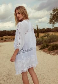 Copertura bianca ricamata con dettagli in pizzo, vestibilità rilassata, maniche a tre quarti e orlo con dettagli decorativi. Tessuto texturizzato.