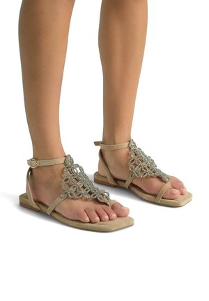 Sandali piatti beige con un design decorativo in perline argento, cinturino alla caviglia e un materiale morbido simile alla pelle scamosciata. Stile open-toe con una texture morbida.