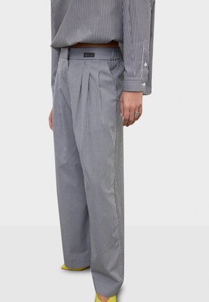 Osoba nosząca luźne, wysokie w talii szare spodnie w kratę z plisami, w paskowaną koszulę, pierścionki i żółte spiczaste buty.