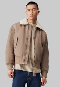 Chaqueta bomber marrón con cuello de borrego, cierre de cremallera, puños de canalé y un bolsillo lateral. Llevada sobre un suéter beige texturizado y unos jeans azules.