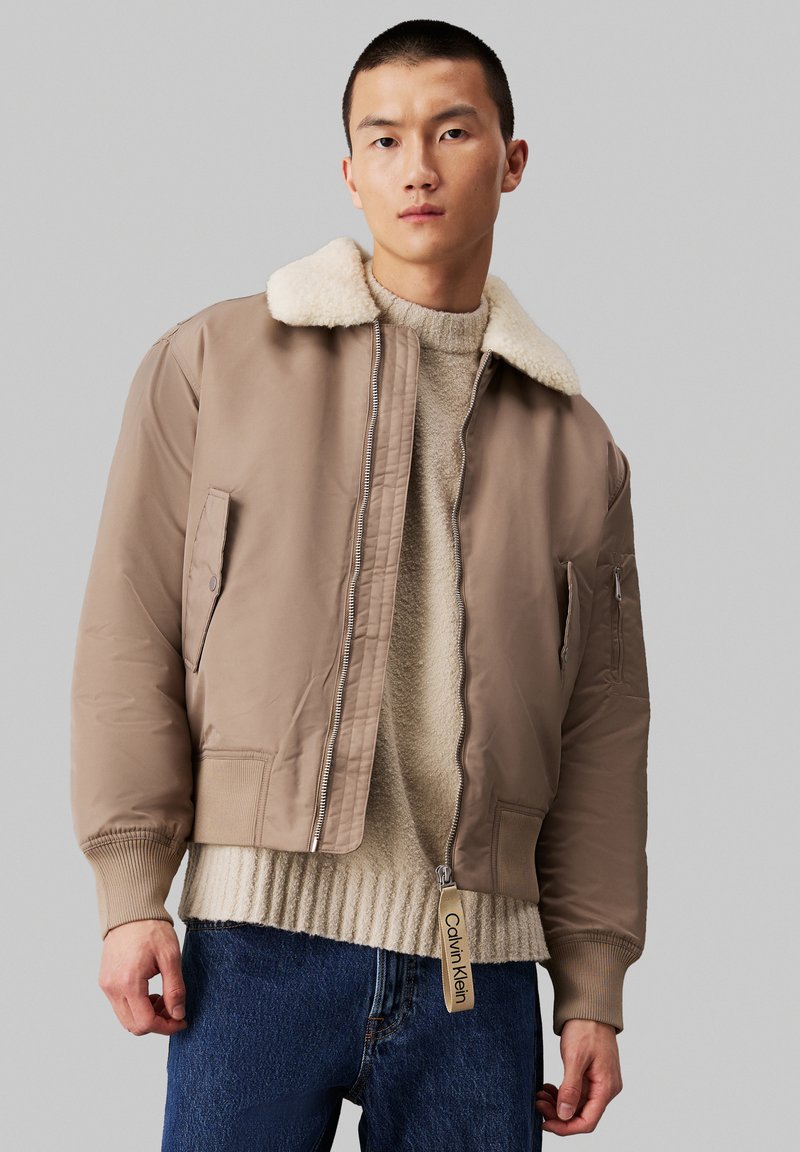 Chaqueta bomber marrón con cuello de borrego, cierre de cremallera, puños de canalé y un bolsillo lateral. Llevada sobre un suéter beige texturizado y unos jeans azules.