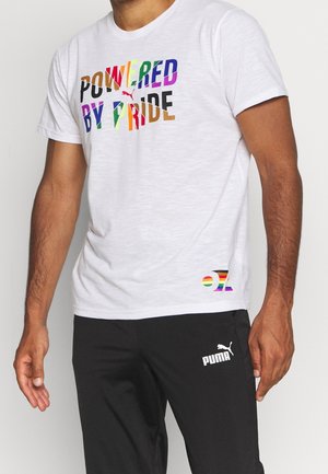 Mężczyzna w białej koszulce z kolorowym napisem "Powered by Pride" i czarnych spodniach Puma na jednolitym tle.