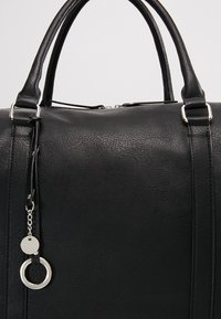 Sort faux læder taske med to runde håndtag, lynlås lukning, tekstureret overflade og sølvfarvet hardware accent med en cirkulær charm.