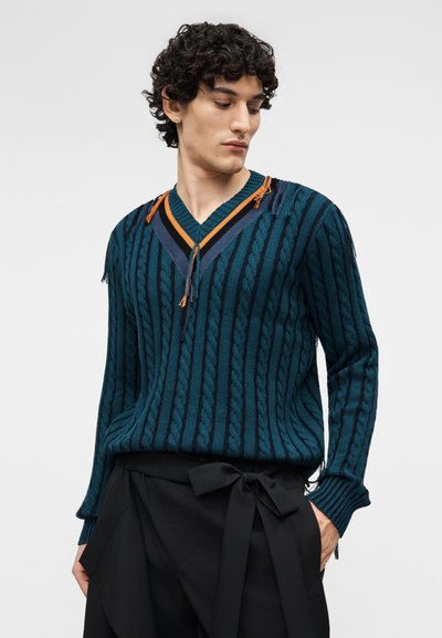 Homme aux cheveux bouclés regardant vers le bas, portant un pull en maille torsadée bleu sarcelle avec un col en V rayé et un pantalon noir avec une ceinture nouée à la taille.