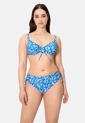 Mujer con bikini azul de estampado floral, con tirantes ajustables y pantaletas con lazos a los lados, de pie contra un fondo claro y liso.