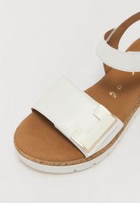 White leather sandal with a wide strap, featuring a gold geometric accent. Cork footbed with a thick white sole and brown textured insole.