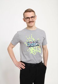 Šedé bavlněné tričko s grafikou v graffiti stylu, zobrazující nákladní auto, a neonově zelenými akcenty, krátké rukávy a kulatý výstřih. Nosené s černými kalhotami.