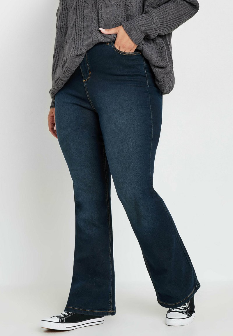 Mörk denim jeans med utsvängda ben och medelhög midja, med gyllene sömmar och en slät yta, tillsammans med svarta canvas-sneakers.