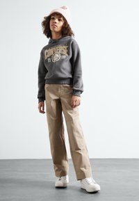 Felpa grigia con grafica "CONVERSE", pantaloni beige con tasche e sneakers bianche. Il modello indossa un berretto rosa chiaro. Sfondo semplice.