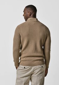 Beige gerippter Pullover mit hohem Kragen, langen Ärmeln und einer taillierten Silhouette, kombiniert mit hellen Hosen mit Gesäßtaschen.