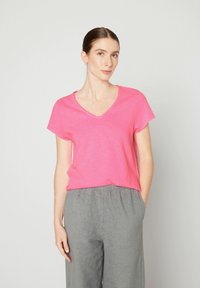 Rožinė medvilninė T-shirt su V iškirpte ir trumpomis rankovėmis, su laisvu kirpimu ir žaliu kraštu, derinama su pilkomis tekstūruotomis kelnėmis.