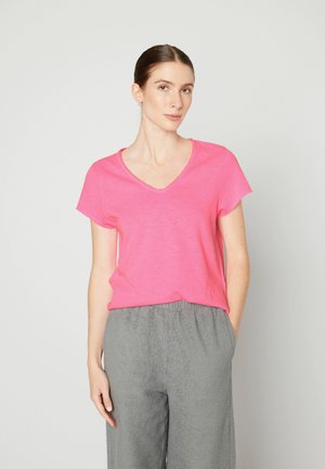 Różowa bawełniana koszulka V-neck z krótkim rękawem, charakteryzująca się luźnym krojem i surowym wykończeniem, zestawiona z szarymi, fakturowanymi spodniami.