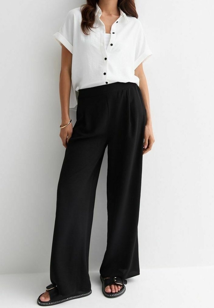 Femme portant une chemise blanche à manches courtes boutonnée, un pantalon noir à jambes larges et des sandales noires avec des détails de boucle.
