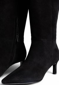 Bottes noires en daim mi-mollet avec des bouts pointus, des talons fins et des zips sur le côté sur fond blanc.