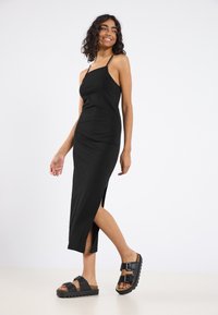 Robe maxi noire ajustée avec des fines bretelles et une fente sur le côté, associée à des sandales noires chunkys avec des doubles boucles et une semelle texturée.