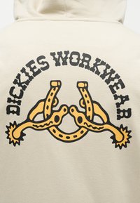 Béžová mikina s kapucňou s čiernym textom "DICKIES WORKWEAR" vo forme oblúka nad dvoma štýlovo žltými ostruhami na chrbte.
