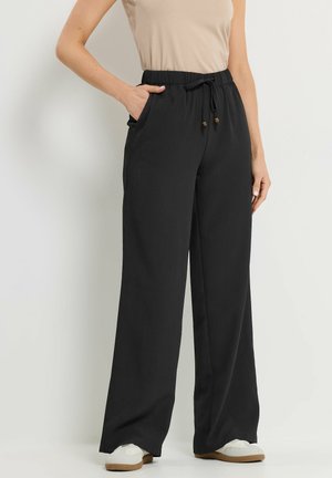 Vrouw draagt zwarte wijde broek met elastische taille en drawstring, beige mouwloze top en witte sneakers, staat met hand in zak.