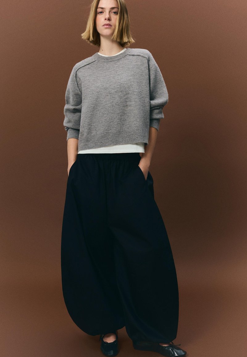 Pulover gri cropped cu mâneci largi, suprapus peste un top alb; asortat cu pantaloni negri largi. Modelul stă cu mâinile în buzunare, fundal neutru.