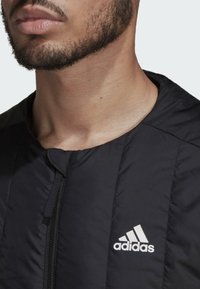 Zwarte gewatteerde jas met een hoge kraag en ritssluiting, voorzien van een wit Adidas-logo op de linkerborst. gladde textuur en strakke lijnen.