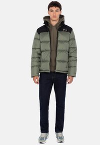 Zielona kurtka typu puffer z czarnym panelem na ramionach, zamkiem błyskawicznym i pikowaną fakturą, noszona na szarej bluzie z kapturem i zestawiona z ciemnymi dżinsami.