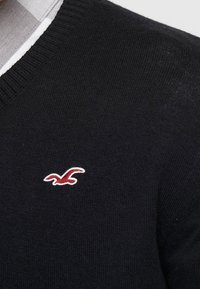 Svart tröja med V-ringning och ribbad krage. Har en liten röd och vit logotyp på vänster bröst. Mjuk stickad textur.